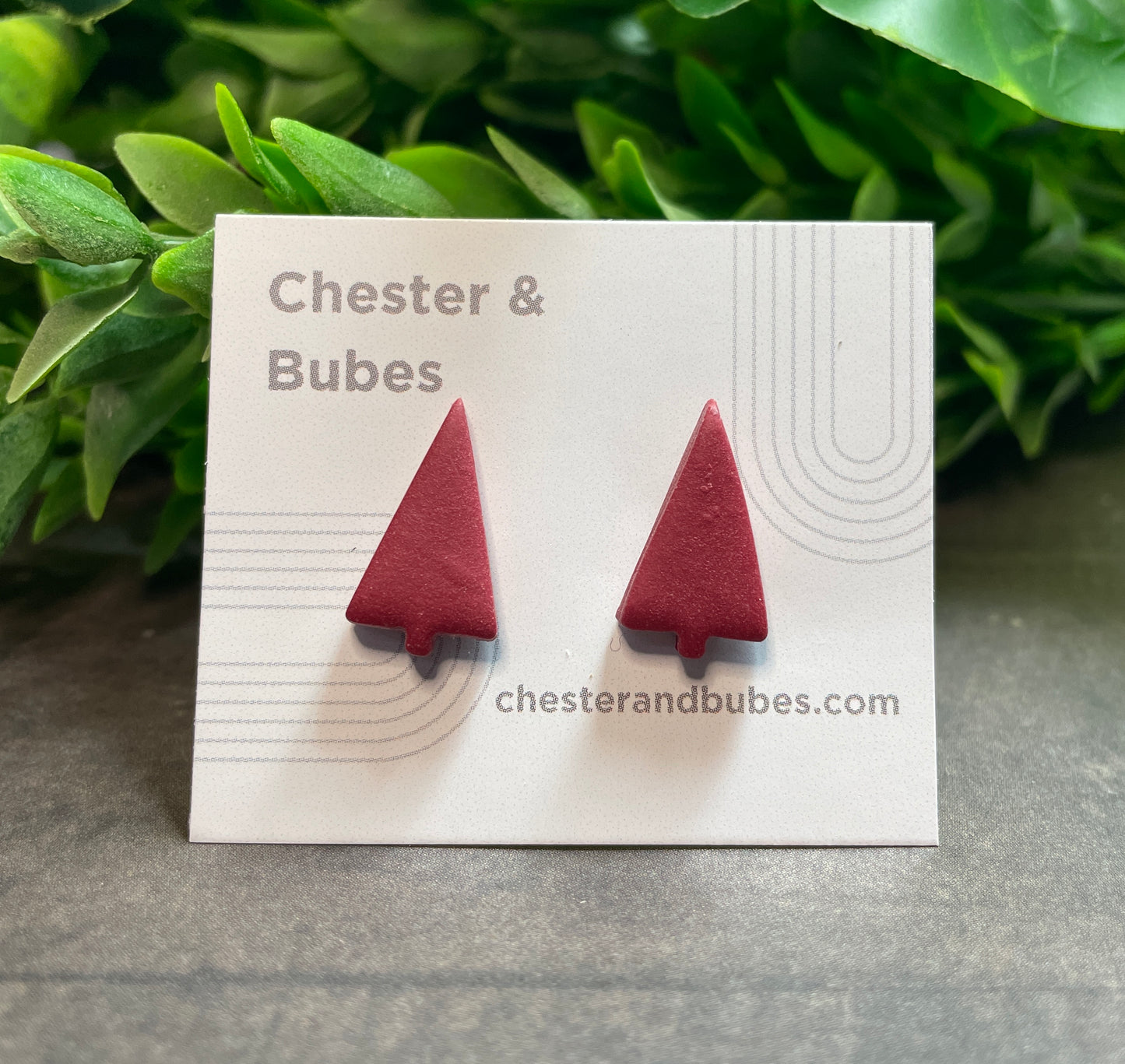 Cranberry red Christmas tree earrings on a 'Chester & Bubes' branded card with green leaves in the background.