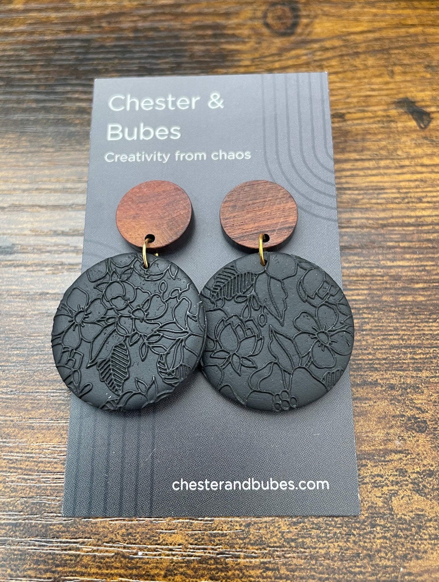 Pair of earrings on a 'Chester & Bubes' branded card with wooden and black designs. Features a detailed floral embossed patter a black clay disc