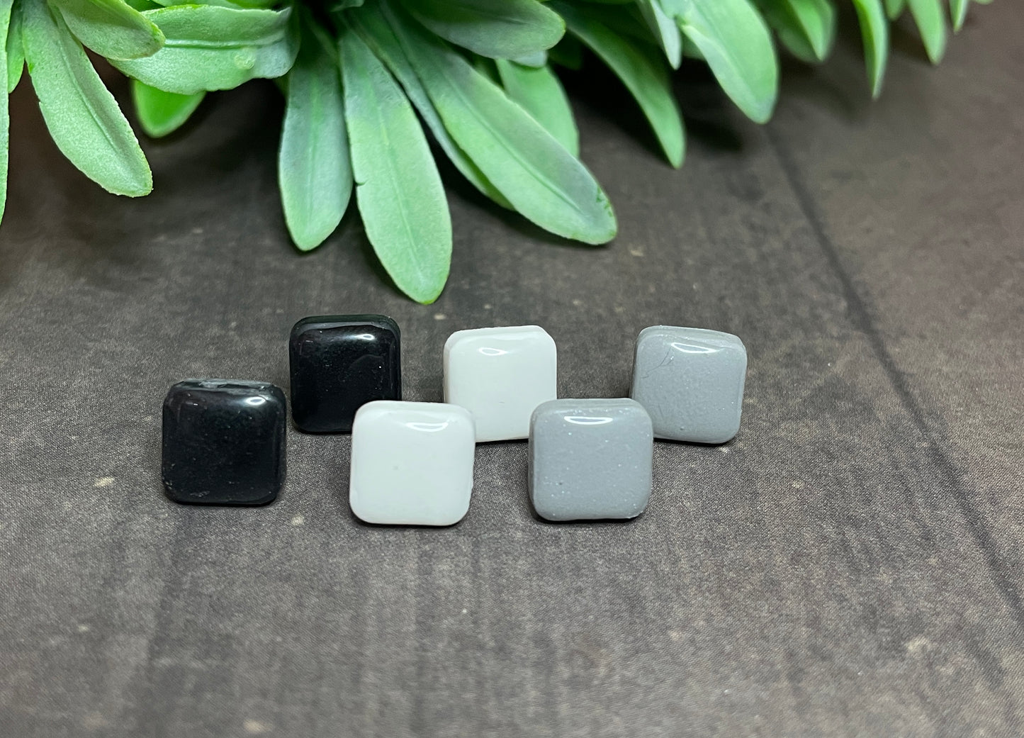 Set of square-shaped earrings in black, white, and gray on a textured surface with green leaves in the background.