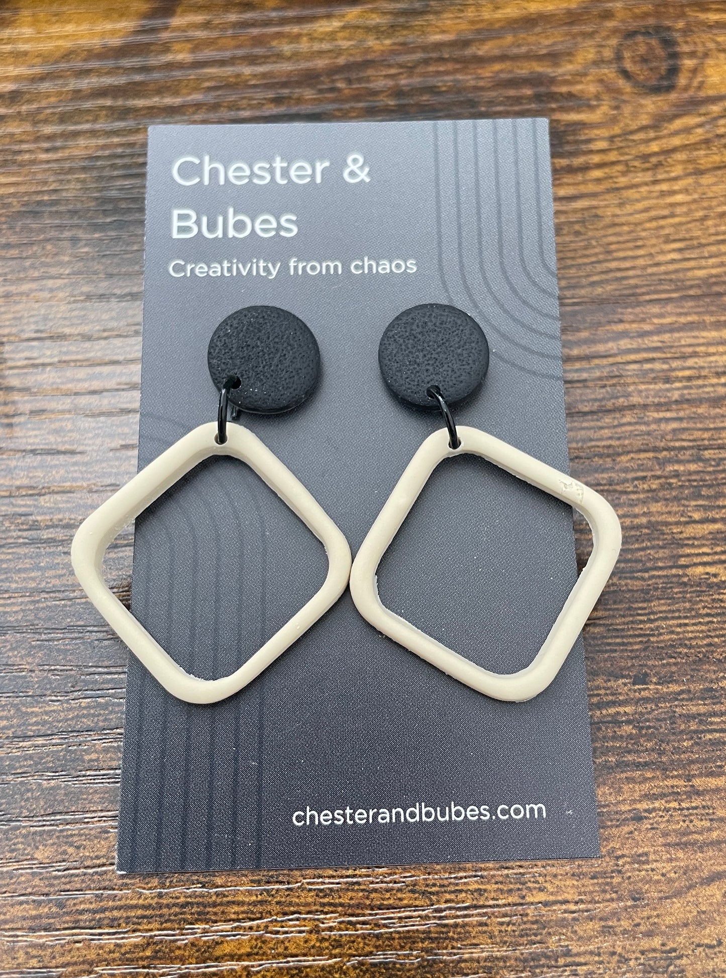 Pair of earrings on a 'Chester & Bubes' branded card. Earrings have a textured black round topper, approx 1.5 cm diameter with a cream coloured offset cutout square.