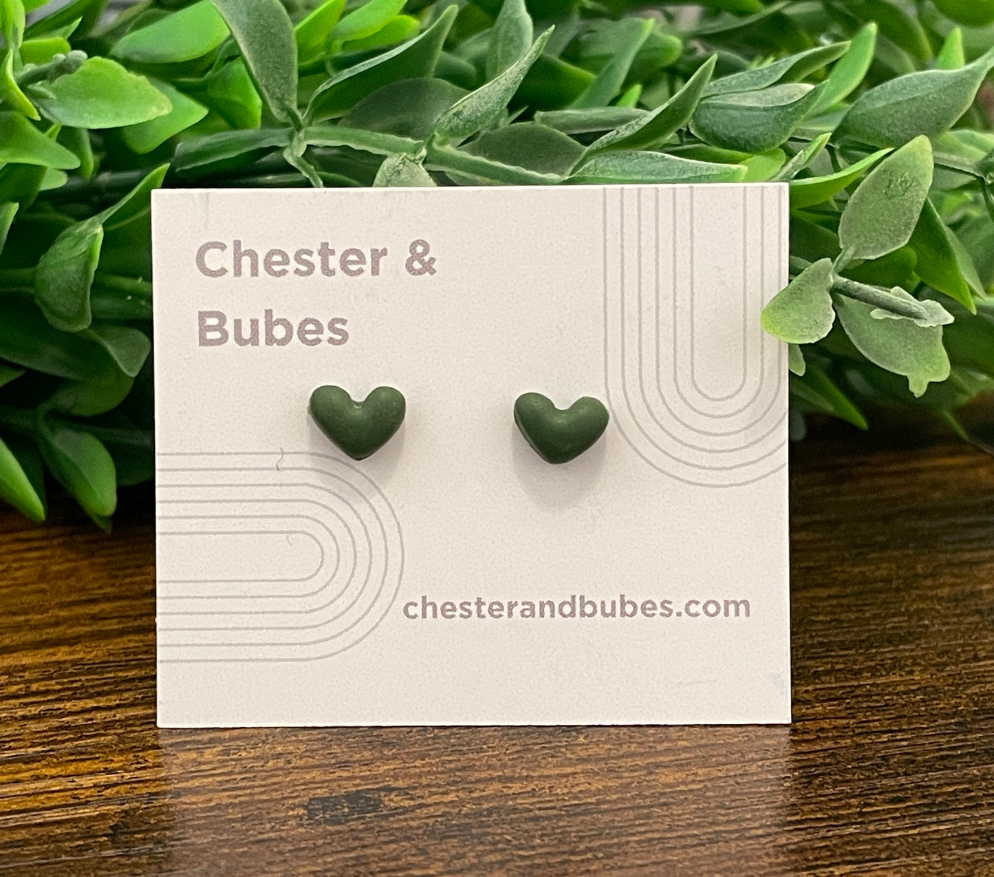 Dark green heart-shaped earrings on a 'Chester & Bubes' branded card with green leaves in the background.