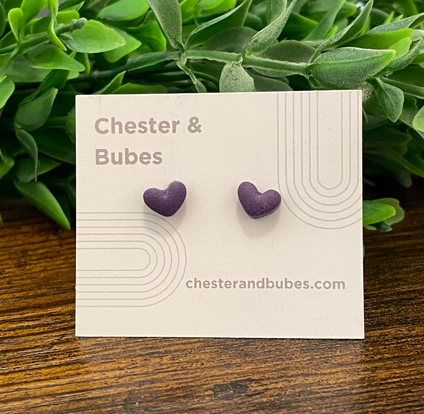 Purple heart-shaped earrings on a Chester & Bubes branded card with green leaves in the background