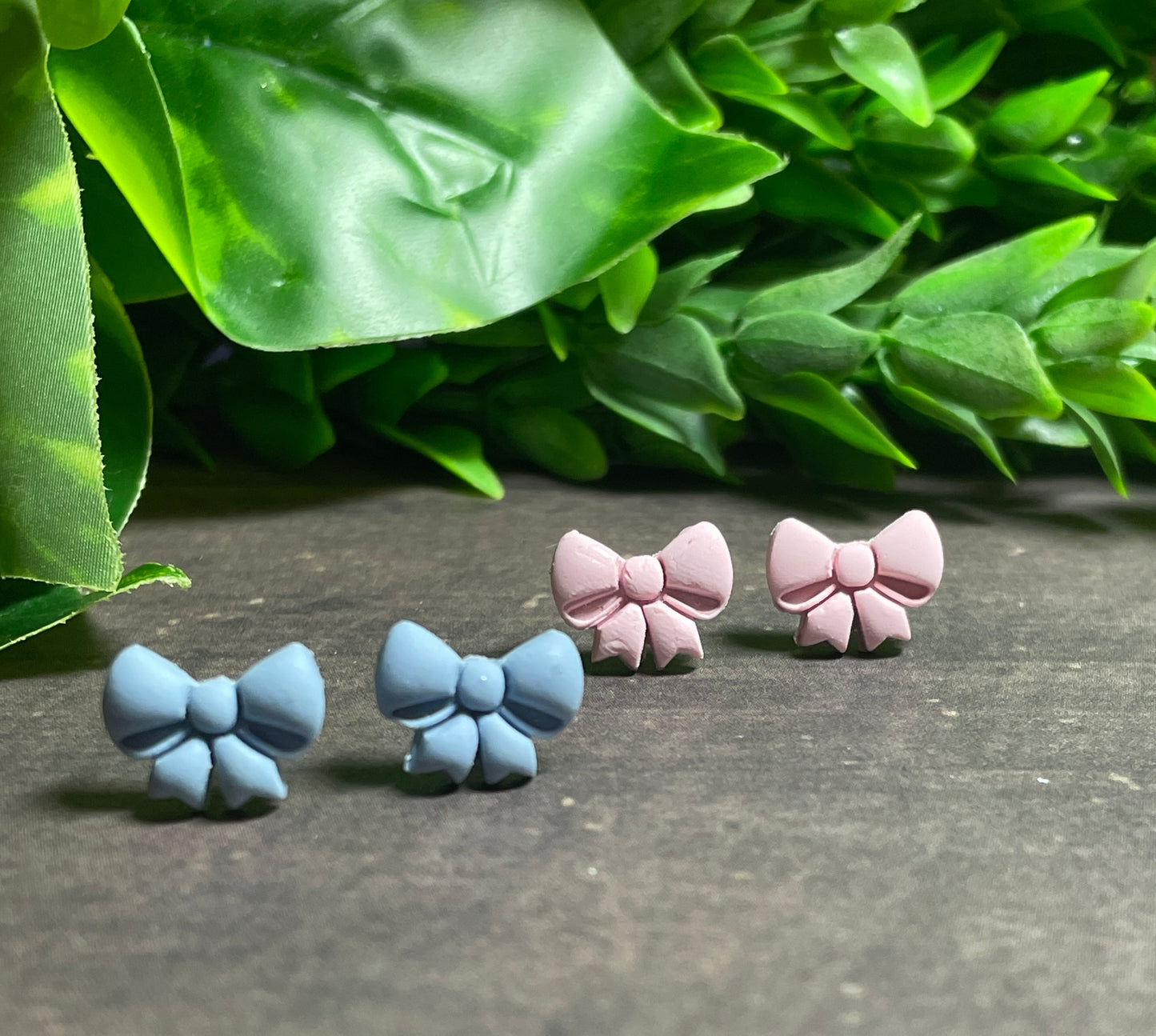 Bow-shaped earrings in blue and pink on a dark surface with green leaves in the background