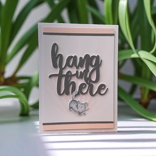 Card with 'hang in there' text and a cat illustration on a light background with green plants in the background