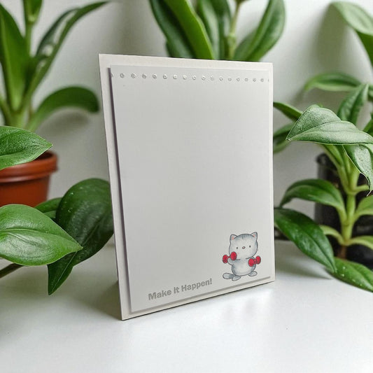 Notebook with a cat illustration and 'Make It Happen!' text, surrounded by green plants.