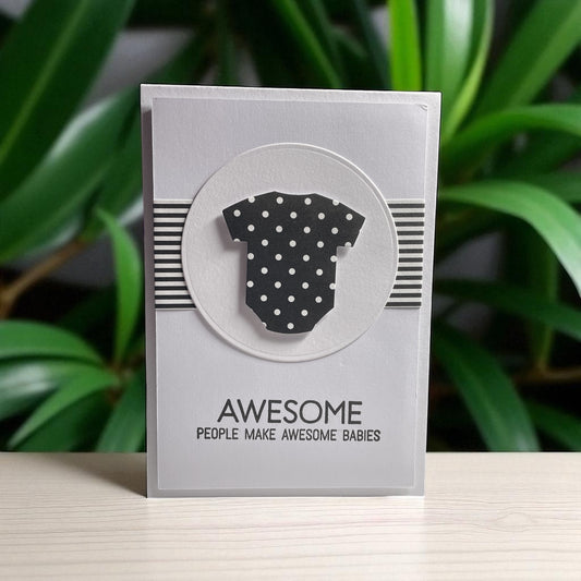 Card with a polka dot baby onesie illustration and text on a gray background with green leaves in the background