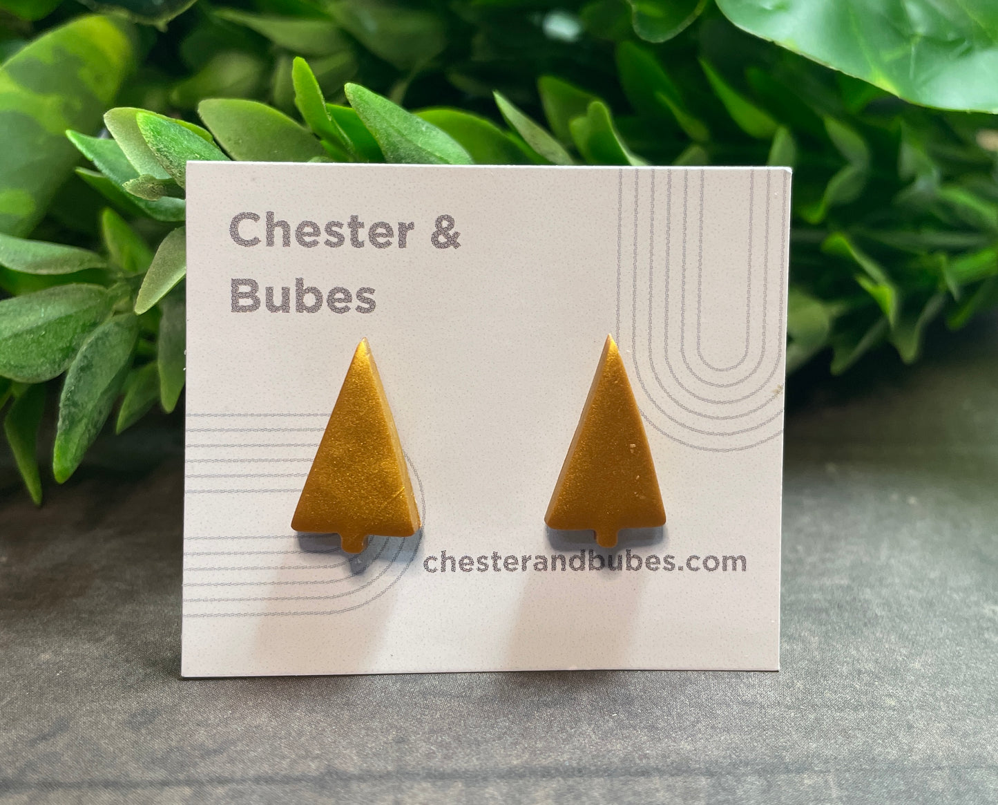 Gold Christmas tree earrings on a 'Chester & Bubes' branded card with green leaves in the background.