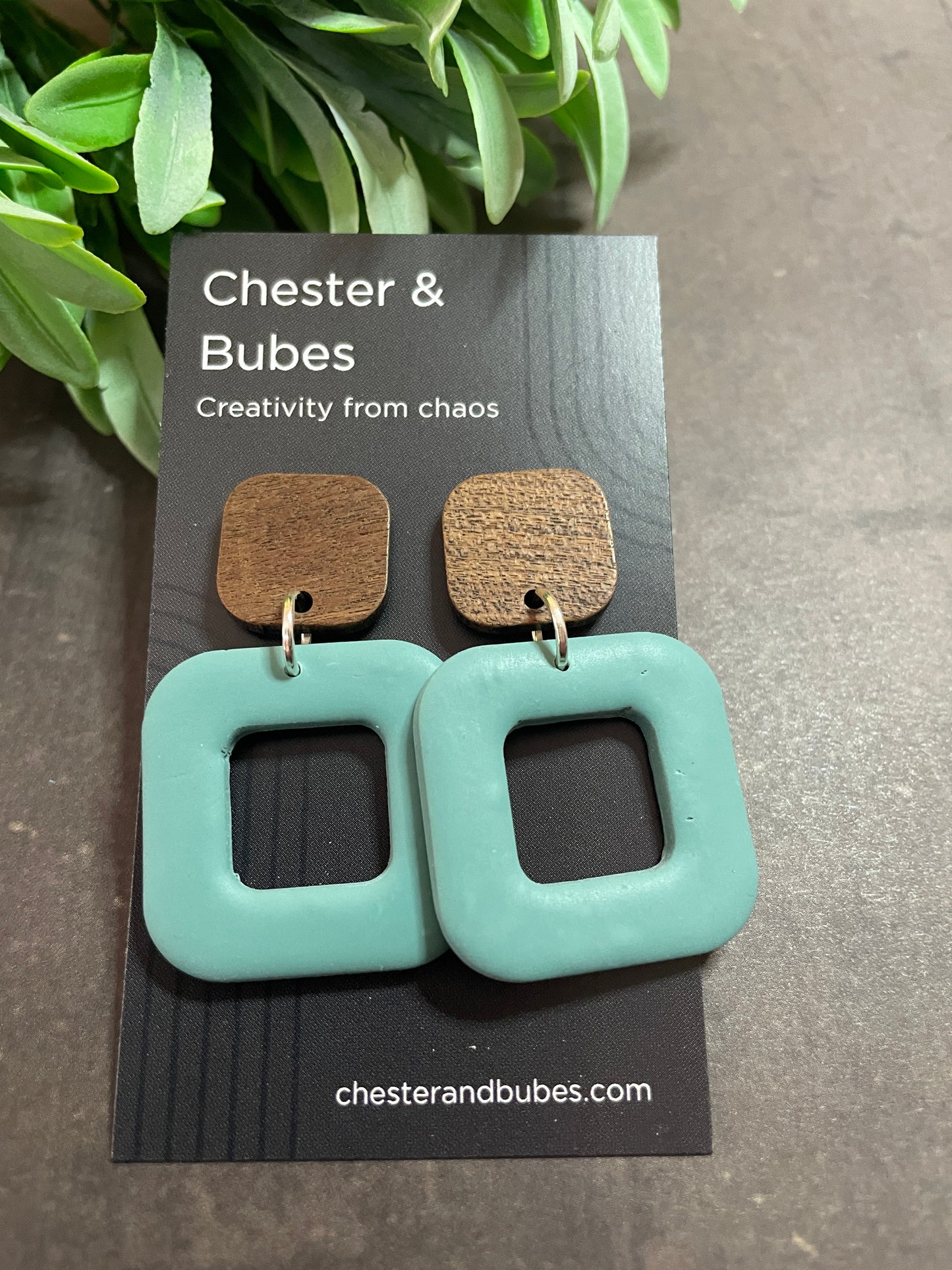 Teal square-shaped earrings on a branded card with a plant in the background