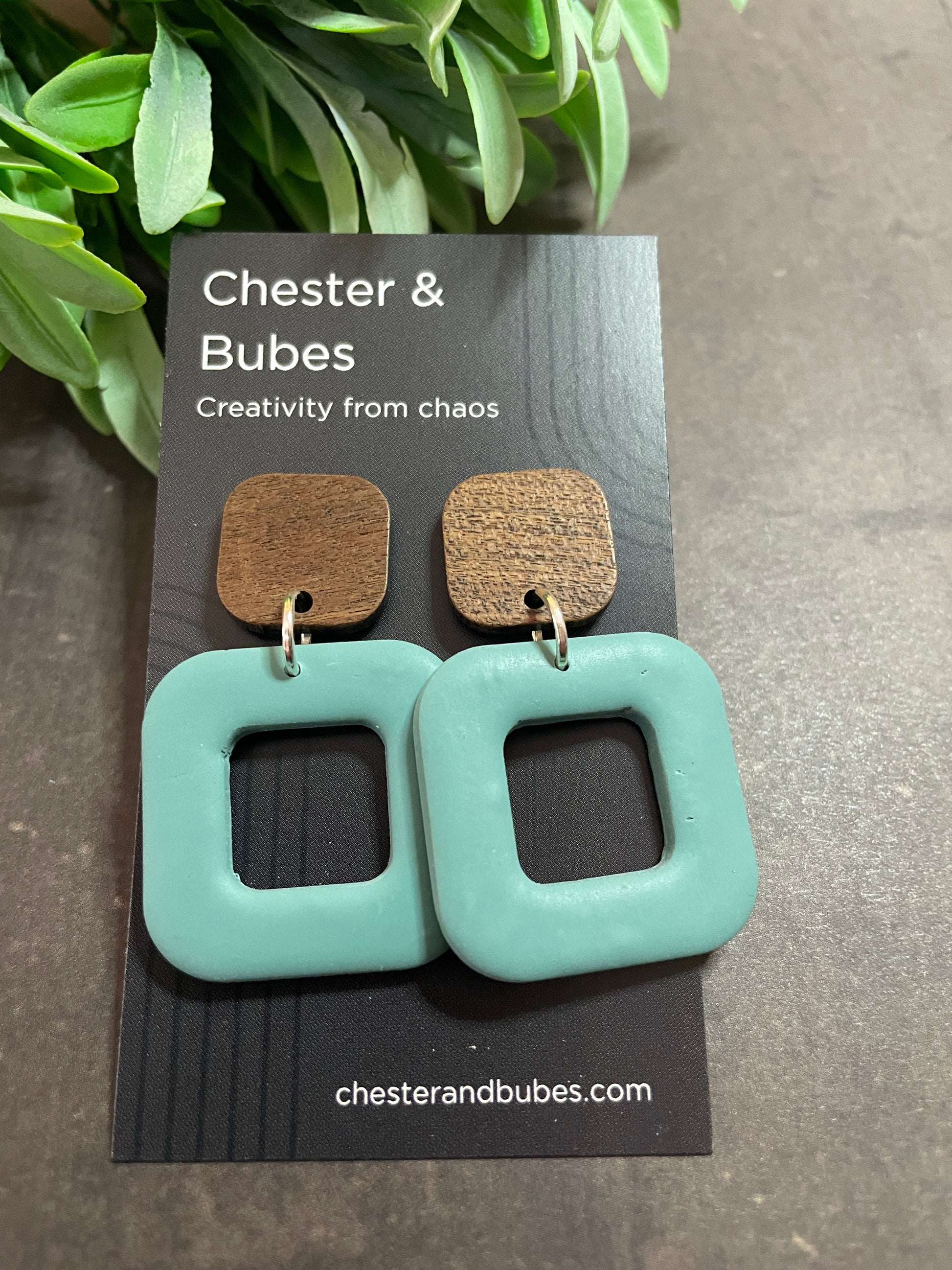 Teal square-shaped earrings on a branded card with a plant in the background